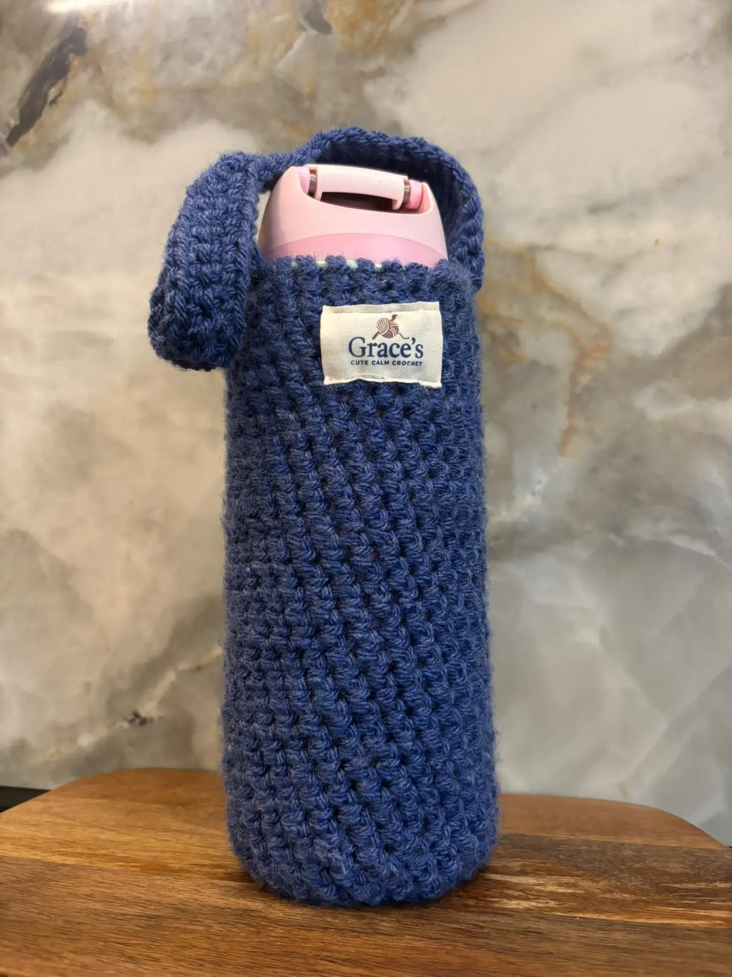 Navy blue crochet bottle carrier with a pink bottle inside and a Grace's Cute Calm Crochet label